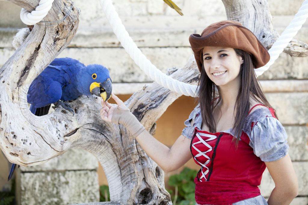 해적 의상을 입은 여성이 파란색과 노란색 앵무새를 들고 미소를 짓고 있습니다. 그녀는 밧줄과 나무 줄기 옆에 서 있습니다.