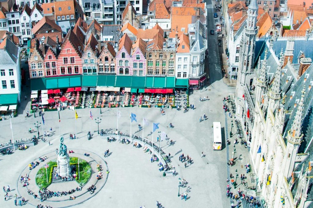 동상, 깃발, 사람들이 있는 크고 분주한 광장의 공중 전망. 빨간색 지붕과 야외 좌석을 배경으로 한 다채로운 건물.