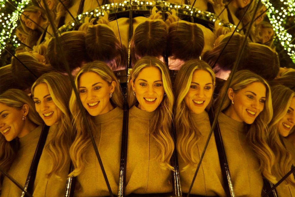 금발의 긴 머리를 한 여성이 따뜻한 조명과 녹색 조명이 보이는 거울에 여러 번 반사됩니다.