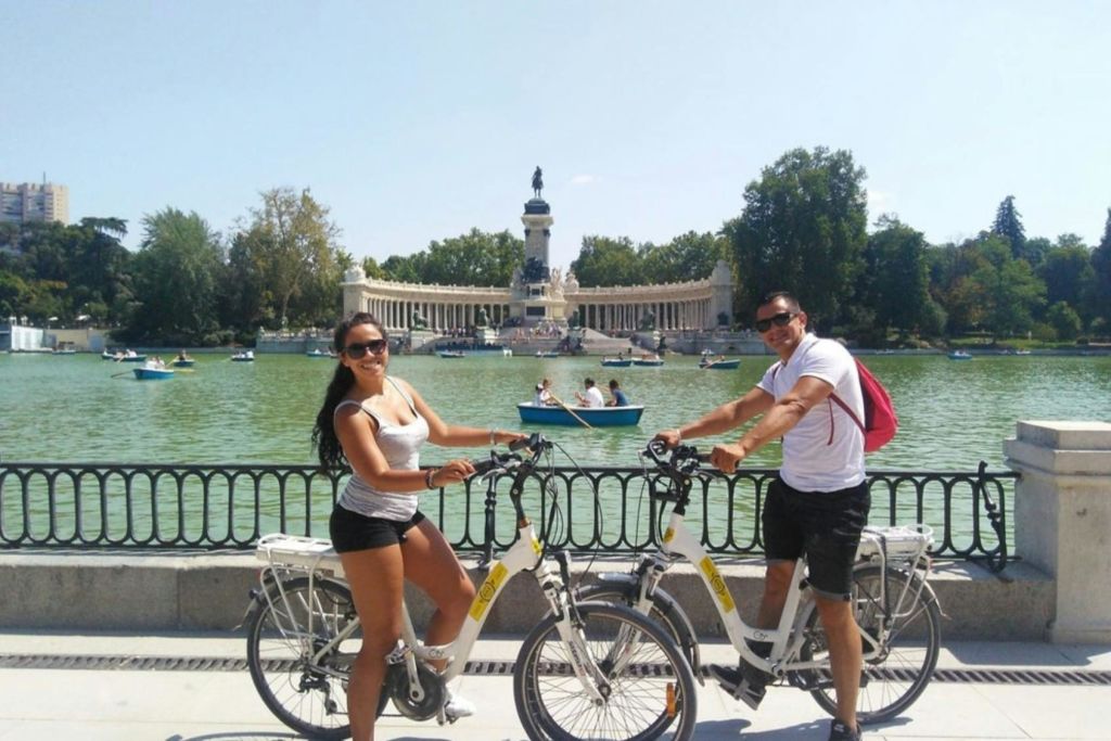 화창한 날 호수 근처에서 자전거를 타고 보트와 기념비, 나무를 배경으로 서 있는 여성.