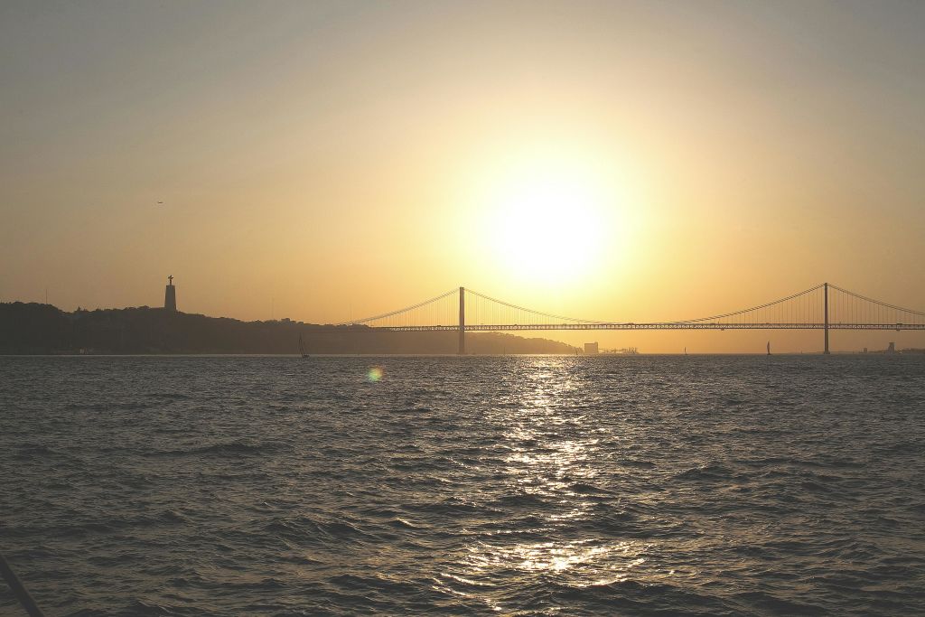 해가 질 무렵 물 위로 현수교가 뻗어 있으며, 왼쪽 수평선에 동상이 있고 머리 위로 햇살이 밝게 비치고 있습니다.