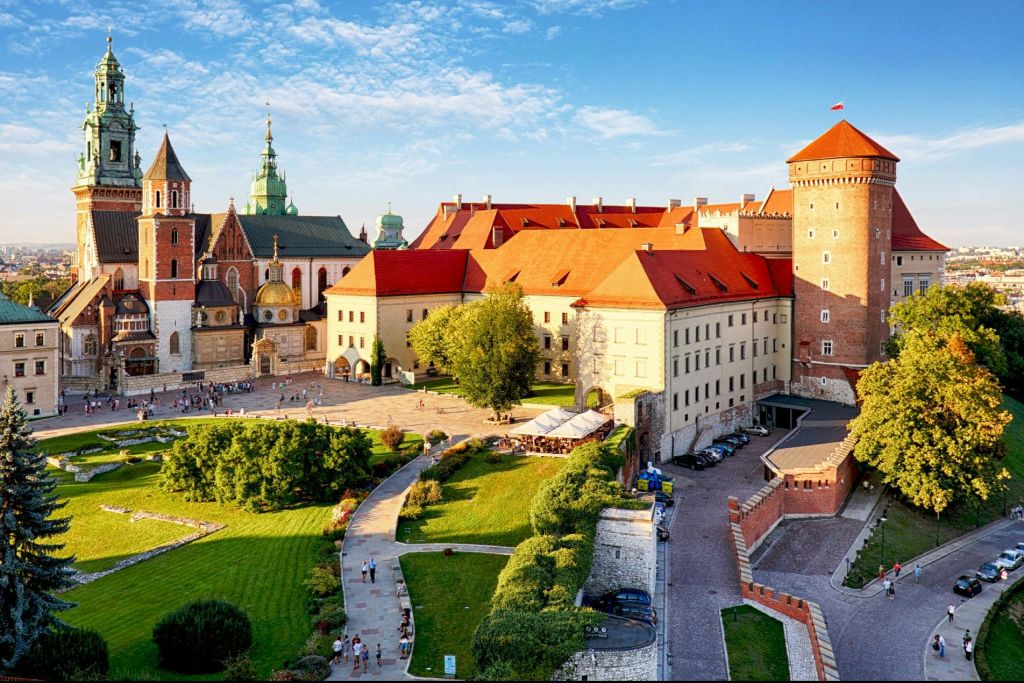 붉은 지붕의 건물과 교회, 푸른 하늘 아래 녹지로 둘러싸인 안뜰이 있는 유서 깊은 유럽 성곽 단지입니다.