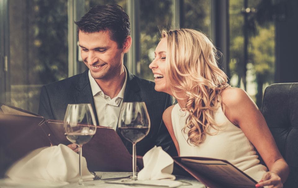 정장을 입은 남성과 흰색 드레스를 입은 여성이 레스토랑 테이블에 앉아 메뉴를 읽으며 미소를 짓고 있습니다. 그 앞에는 와인잔 두 개가 놓여 있습니다.