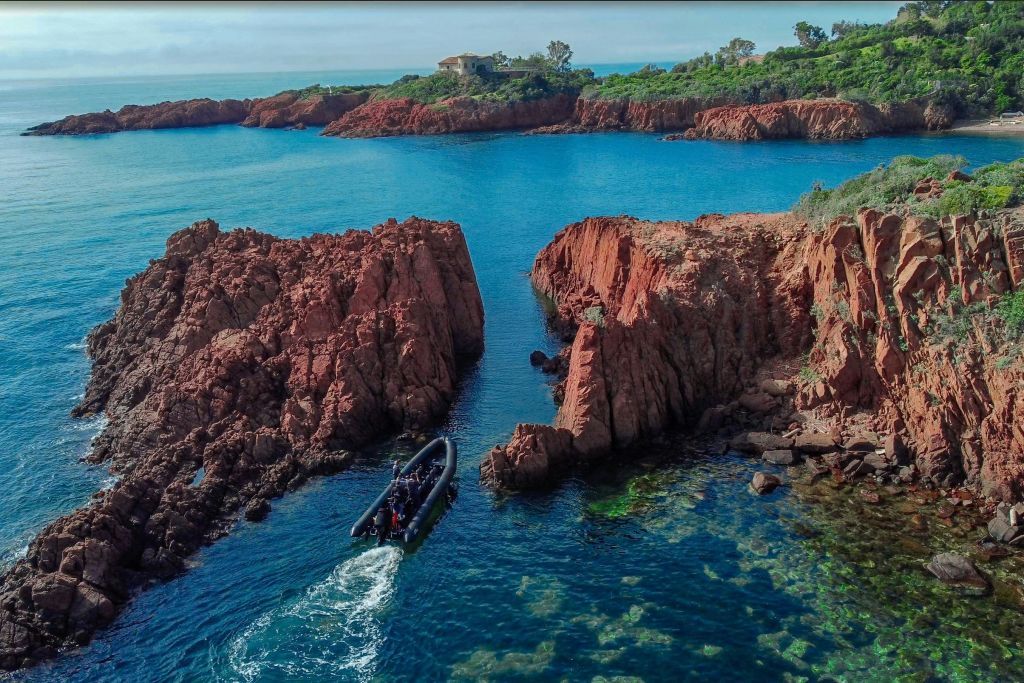 푸른 바다의 붉은 바위 지층 사이를 항해하는 작은 보트로, 먼 해안선과 녹지를 배경으로 합니다.