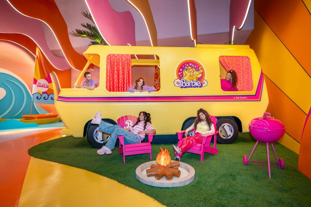 노란색 캠핑카, 분홍색 의자, 모닥불, 초록색 인조 잔디에 분홍색 그릴이 있는 바비를 테마로 한 세팅을 즐기는 네 명.