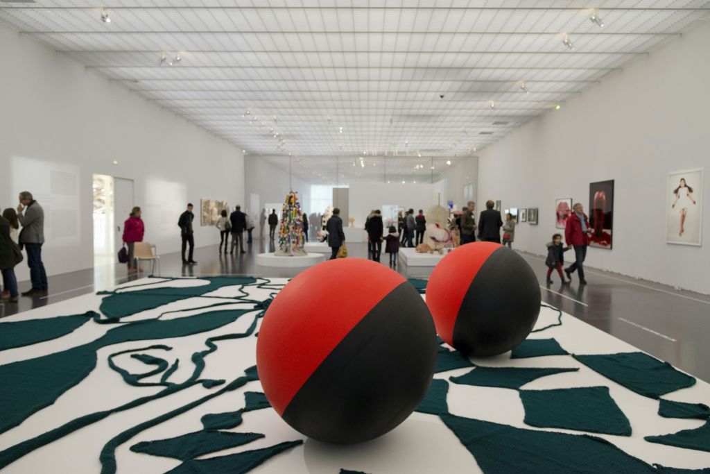 빨간색과 검은색의 커다란 구체와 흰색 벽에 다양한 예술품이 전시된 넓은 아트 갤러리를 둘러보는 사람들.