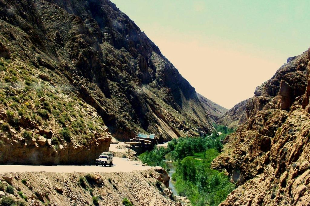 좁은 도로가 건조한 산을 휘감고, 흰색 차량이 도로변에 주차되어 있으며, 푸른 단풍과 아래에 작은 개울이 있습니다.