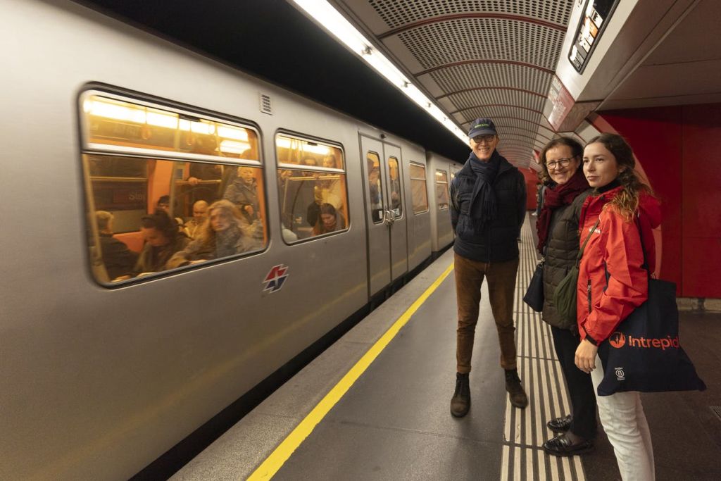 정차 열차 옆의 지하철 플랫폼에 세 사람이 서 있으며, 내부에는 눈에 띄는 승객이 있고 배경에는 빨간색 벽이 있습니다.