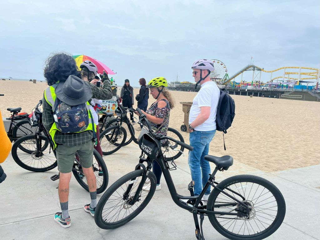헬멧과 자전거를 착용한 사람들이 해변 길에 서 있으며, 배경에는 롤러코스터가 보입니다.