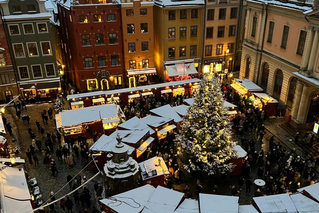 조명이 켜진 가판대, 커다란 장식 트리, 눈 덮인 옥상이 사람들로 붐비는 축제 분위기의 크리스마스 마켓입니다.