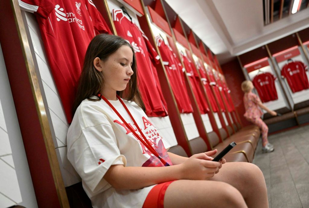 소녀는 라커룸의 벤치에 앉아 휴대전화를 바라보고 있습니다. 빨간색 축구 유니폼이 배경에 걸려 있습니다.