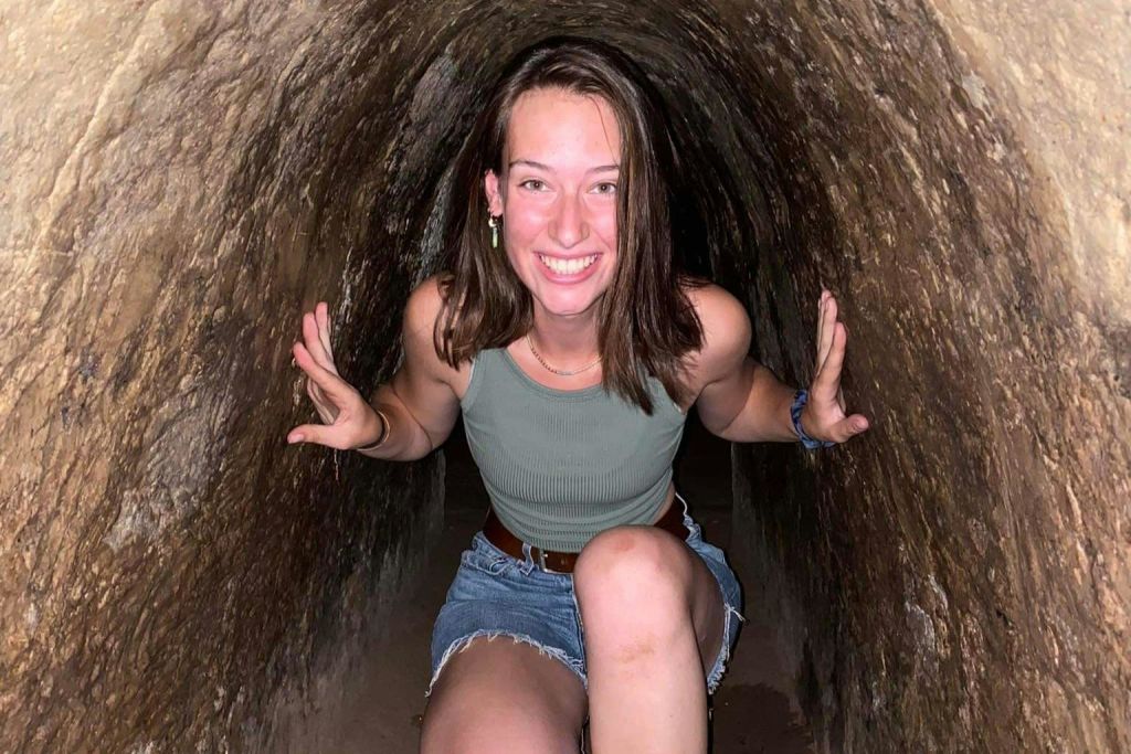 거칠고 질감이 있는 벽이 있는 좁고 원통형의 터널 안에서 한 사람이 웅크리고 미소를 짓고 있습니다. 녹색 탱크탑과 데님 반바지를 입고 있습니다.