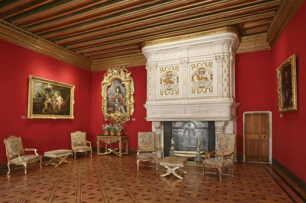 붉은 벽, 화려한 천장, 커다란 장식 벽난로, 앤티크 가구, 액자에 담긴 예술품이 있는 우아한 객실입니다.