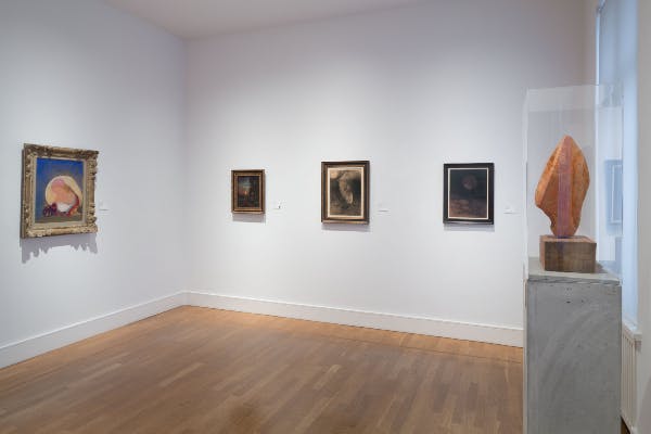나무 바닥이 있는 갤러리의 흰색 벽에 세 개의 액자 그림이 있습니다. 오른쪽에 사람의 팔이 부분적으로 보입니다.