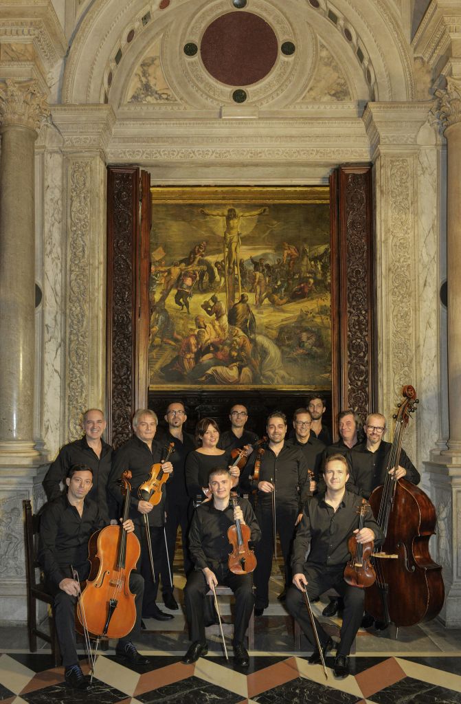 검은색 옷을 입고 현악기를 들고 있는 11명의 사람들이 기둥과 커다란 그림이 있는 화려한 방에서 포즈를 취하고 있습니다.