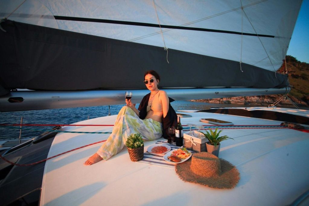 한 사람이 음식 접시, 모자, 화분으로 둘러싸인 요트 데크에 앉아 와인 한 잔을 마시고 있습니다.