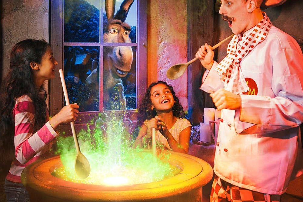 두 소녀와 한 남자가 초록빛을 내뿜는 가마솥을 휘젓고, 만화 당나귀가 뒤에서 창문을 들여다봅니다.