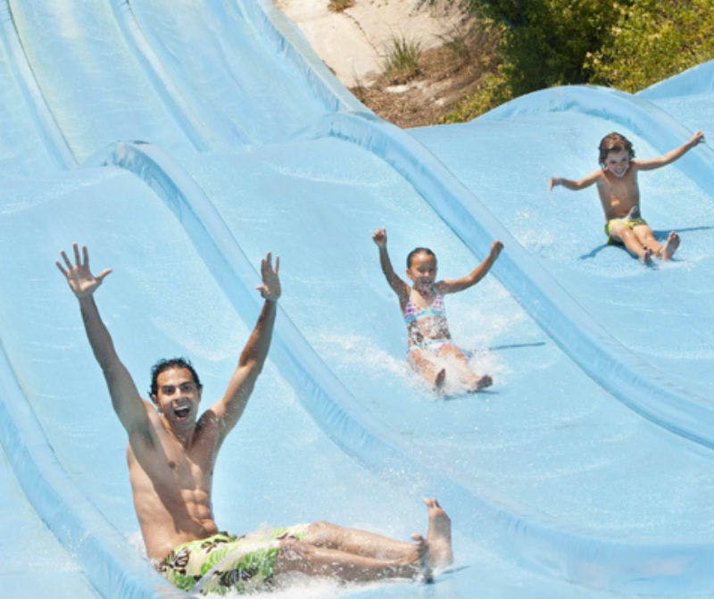 성인 한 명과 어린이 두 명을 포함한 세 명이 팔을 들고 웃는 표정으로 평행 워터 슬라이드를 아래로 미끄러집니다.