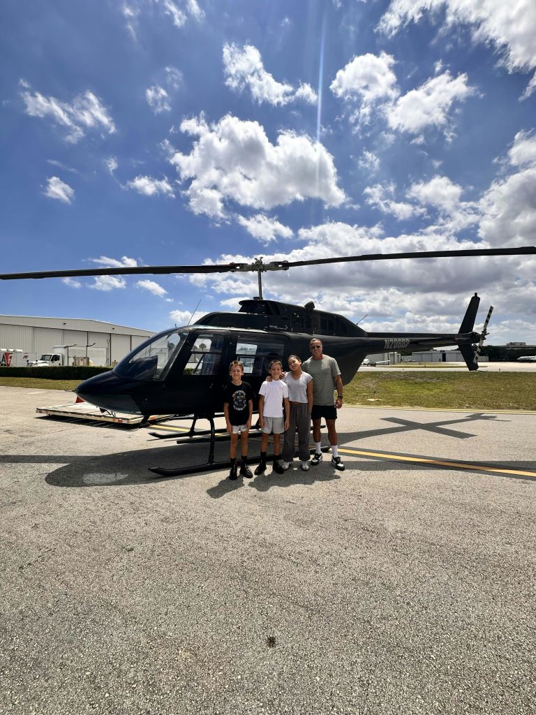 건물과 부분적으로 흐린 하늘을 배경으로 비행장에 주차된 헬리콥터 앞에 서 있는 네 사람.