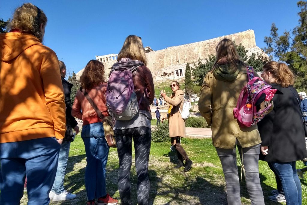 녹음이 우거진 화창한 날, 파르테논 신전 앞에서 한 무리의 관광객과 이야기를 나누는 가이드.