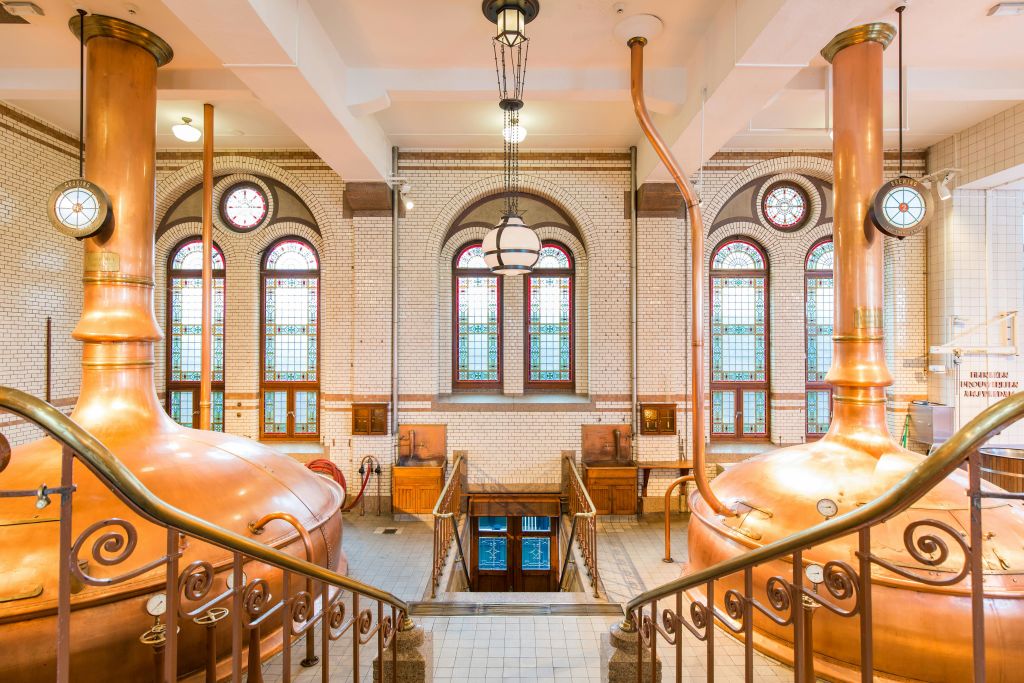 커다란 구리 주전자, 화려한 창문, 아래 문으로 이어지는 계단이 있는 밝은 양조장 내부.