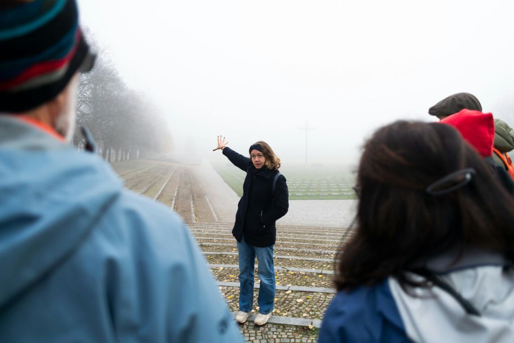 검은 재킷을 입은 사람이 안개가 자욱한 야외 환경에서 한 손으로 계단에 서서 제스처를 취하고 있으며, 멀리 십자가가 있습니다.