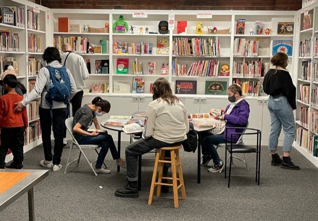 책, 피규어, 다채로운 색상의 물품이 가득한 선반이 있는 도서관에서 책을 읽고 탐색하는 사람들.