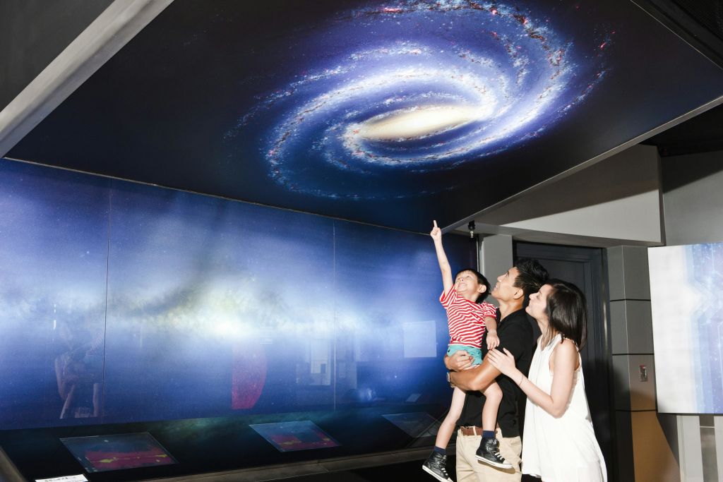 한 가족이 전시관에서 크고 상세한 은하계 디스플레이를 올려다보고, 한 어린이가 소용돌이치는 불빛을 흥미롭게 가리킵니다.