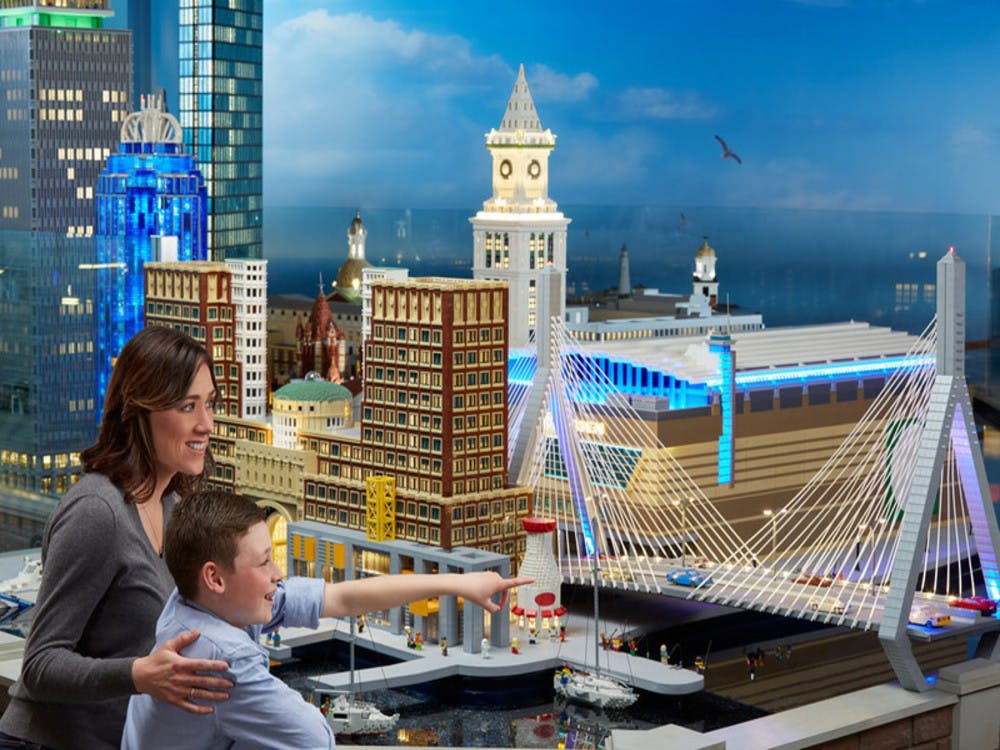 높은 건물, 시계탑, 푸른 하늘 아래 다리가 있는 상세한 레고 도시 풍경에 한 여성과 어린이가 감탄합니다.