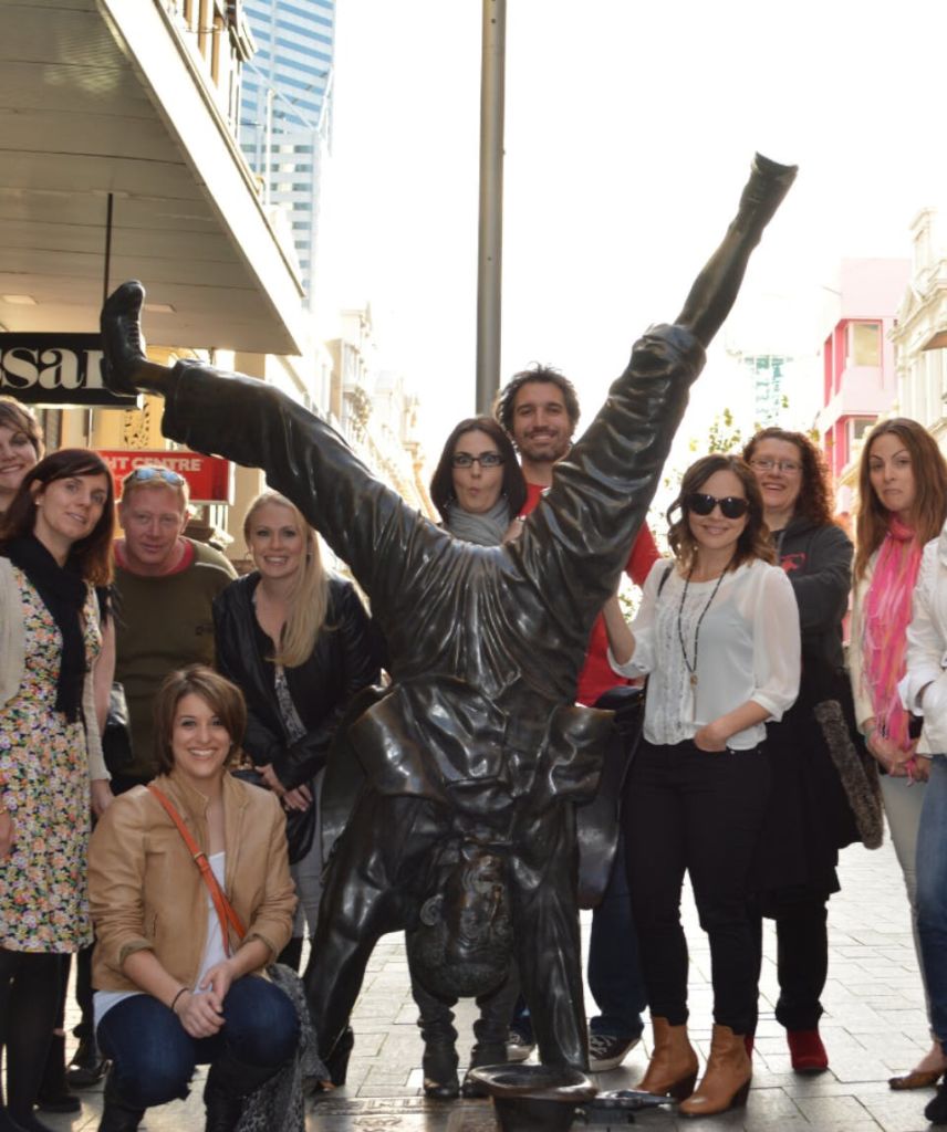 건물과 상점 표지판을 배경으로 도시의 거꾸로 된 동상 주위에 포즈를 취하는 사람들의 그룹.