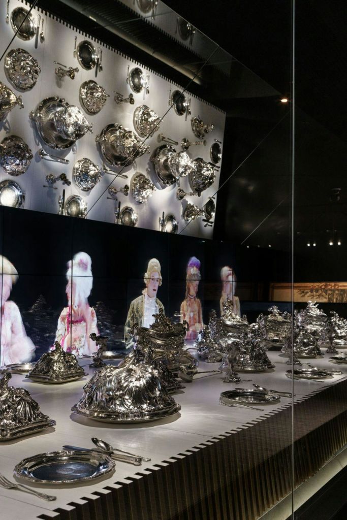 선반과 벽에 다양한 은제품이 진열되어 있고, 역사적인 복장을 한 사람들의 홀로그램이 배경으로 진열되어 있습니다.