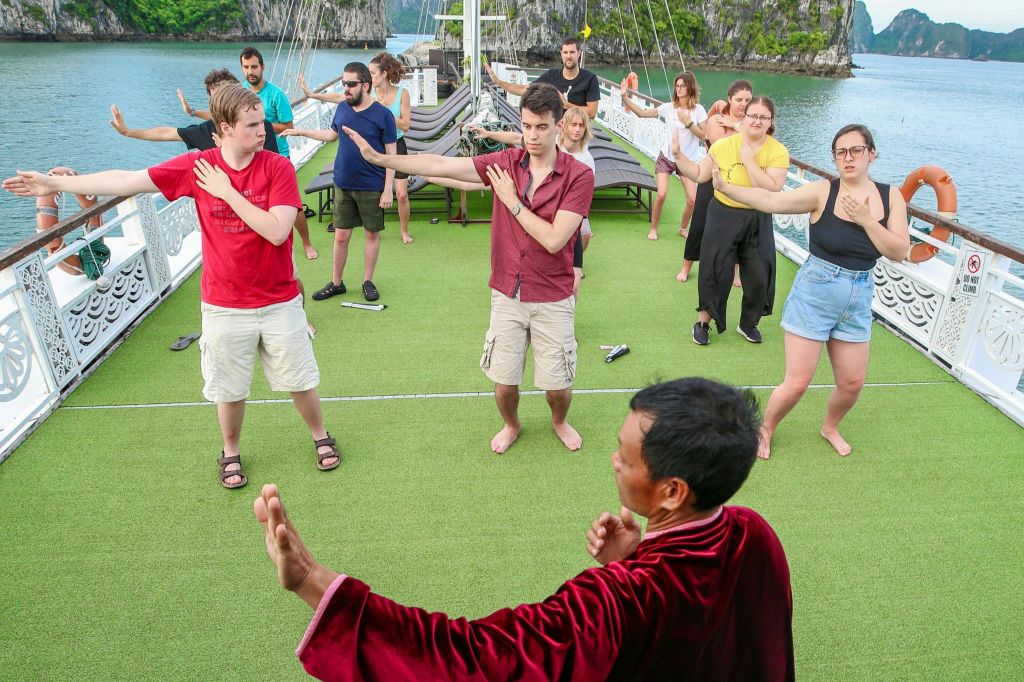 빨간 가운을 입고 바위 절벽을 배경으로 강사가 이끄는 보트 갑판에서 태극권을 연습하는 사람들입니다.
