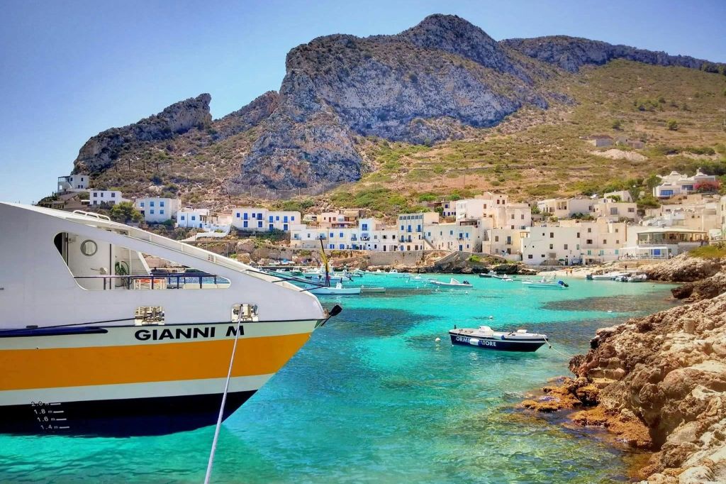 하얀색 건물과 푸른색으로 포인트를 준 해안 마을, 청록색의 맑은 물, 보트, 바위 언덕이 배경으로 펼쳐집니다.