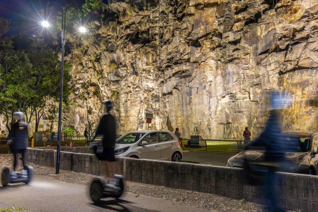 밤에 불빛이 비추는 암벽 등반, 등반하는 사람, 근처에 주차된 차, 전경에 있는 스쿠터에 탄 사람.