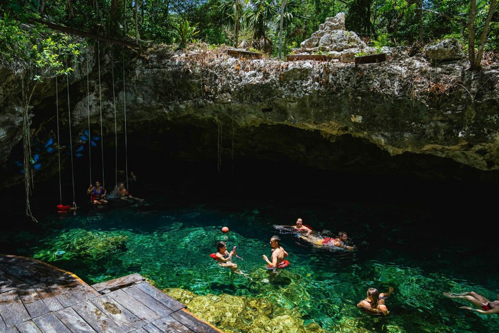 바위 절벽과 초목으로 둘러싸인 맑은 물 동굴 수영장에서 수영하고 휴식을 취하는 사람들.