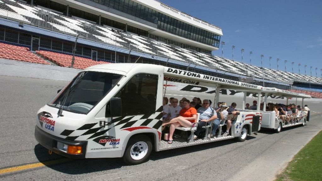 '데이토나 인터내셔널 스피드웨이 (Daytona International Speedway)' 라는 이름의 트램을 타는 한 무리의 사람들이 그랜드스탠드를 배경으로 서 있습니다.
