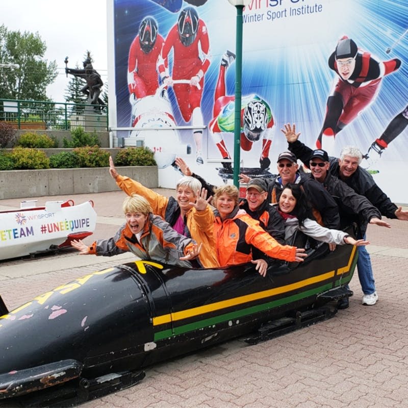 8명의 사람들이 검은 봅슬레이 안에 앉아 겨울 스포츠 선수들이 그려진 벽화 앞에서 두 팔을 번쩍 들고 포즈를 취하고 있습니다.