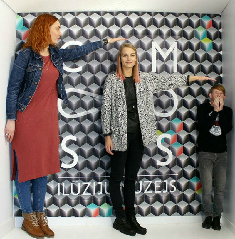 착시 효과가 있는 방에 세 사람이 서 있습니다. 배경에는 기하학적 패턴과 부분적으로 보이는 텍스트가 있습니다.