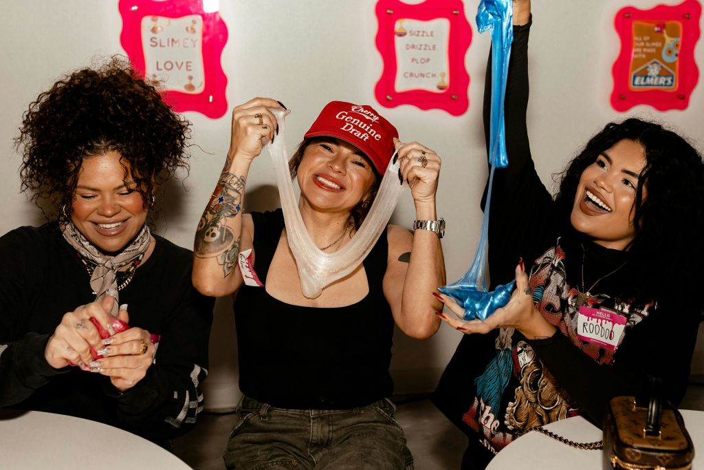 두 여성이 웃으며 슬라임을 가지고 놀고 있습니다. 한 여성은 빨간색 모자를 쓰고, 다른 여성은 스카프를 착용합니다. 배경에 텍스트가 있는 밝은 표지판.