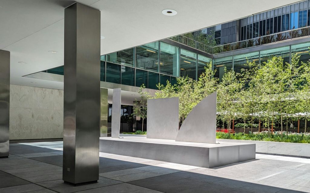 지붕 아래 금속 조각품, 나무, 유리 건물 정면이 있는 현대적인 야외 광장입니다.