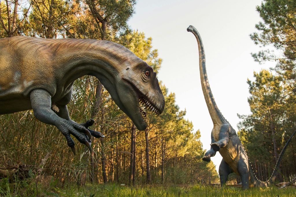 나무가 우거진 지역에 있는 두 개의 살아있는 공룡 조각상. 하나는 목이 길고 다른 하나는 이빨이 날카로우며 나무와 풀로 둘러싸여 있습니다.