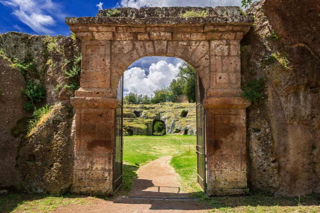 바위 벽으로 둘러싸인 고대 유적과 부분적으로 흐린 하늘 아래 잔디가 우거진 지역으로 이어지는 석조 아치길.