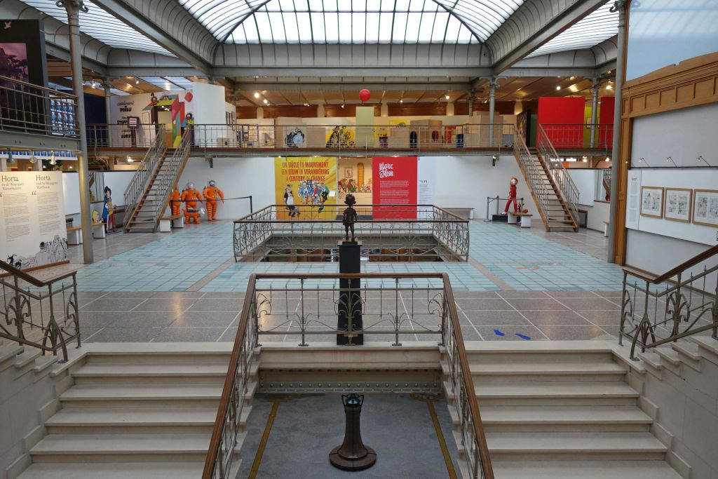 중앙 동상, 갤러리로 이어지는 계단, 다채로운 전시물, 주황색 유니폼을 입은 마네킹이 특징인 박물관 내부.