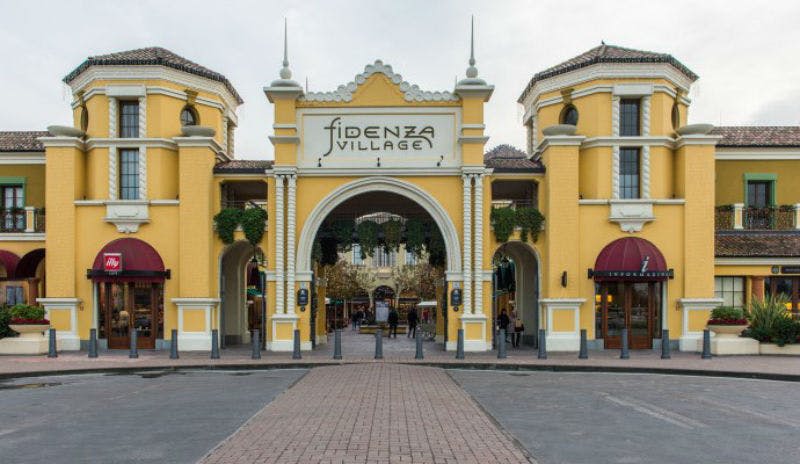 "피덴자 빌리지" 라고 표시된 노란색 아치길 입구 양쪽에 상점과 자갈길이 있습니다.