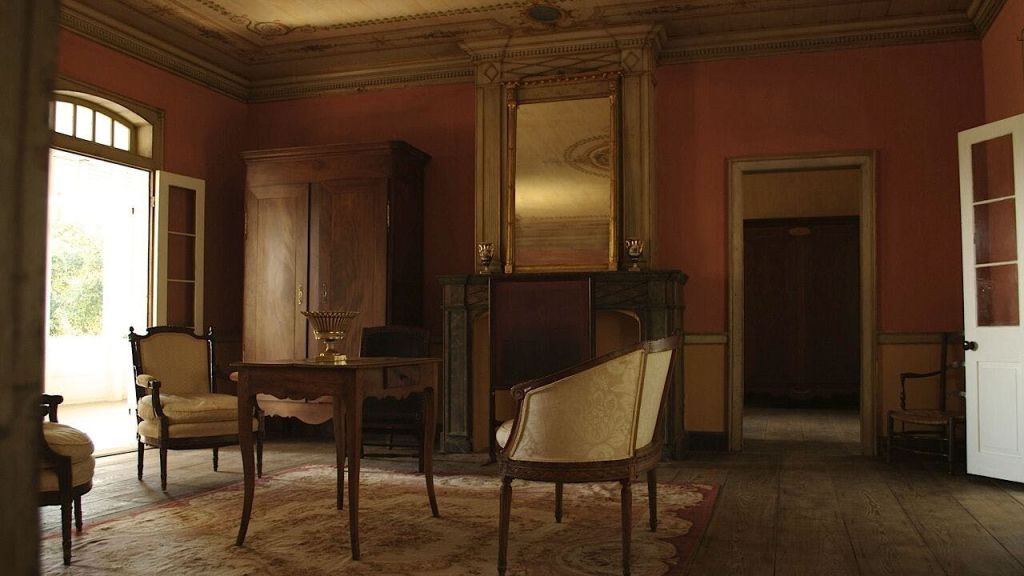 나무 테이블, 조각된 의자, 찬장, 벽난로 위의 높은 거울, 화려한 천장 디테일이 있는 빈티지 객실입니다.