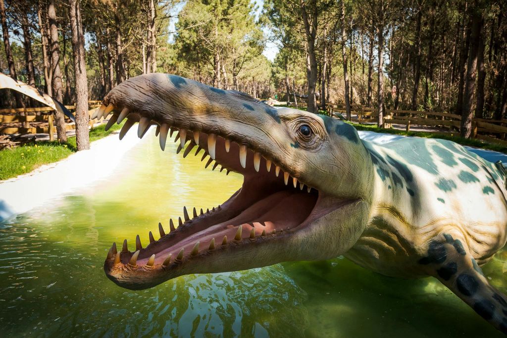 나무로 둘러싸인 녹색의 물이 가득한 지역에서 입을 벌리고 날카로운 이빨을 가진 실물 크기의 공룡 모델.