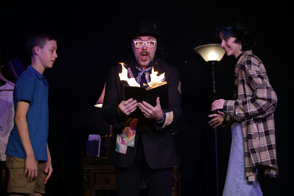 모자와 안경을 쓴 남성이 불타는 책을 들고 있고, 파란색 셔츠와 반바지를 입은 소년이 희미하게 빛나는 무대에서 시계를 보고 있습니다.