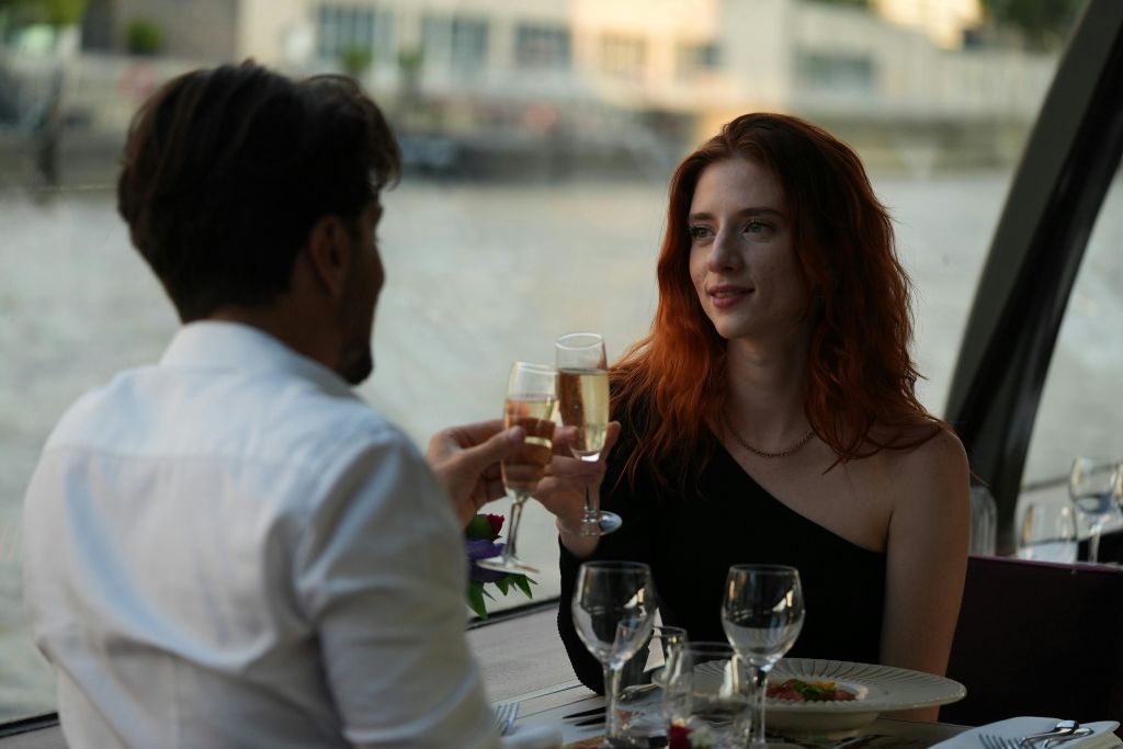 검은 머리를 한 여성이 와인잔이 놓인 식탁에 앉아 다른 사람과 함께 토스트를 하고 있습니다.