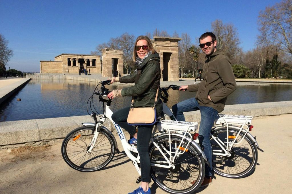 자전거를 탄 두 사람이 반사 수영장과 맑고 푸른 하늘을 배경으로 데보드 사원 앞에서 포즈를 취하고 있습니다.