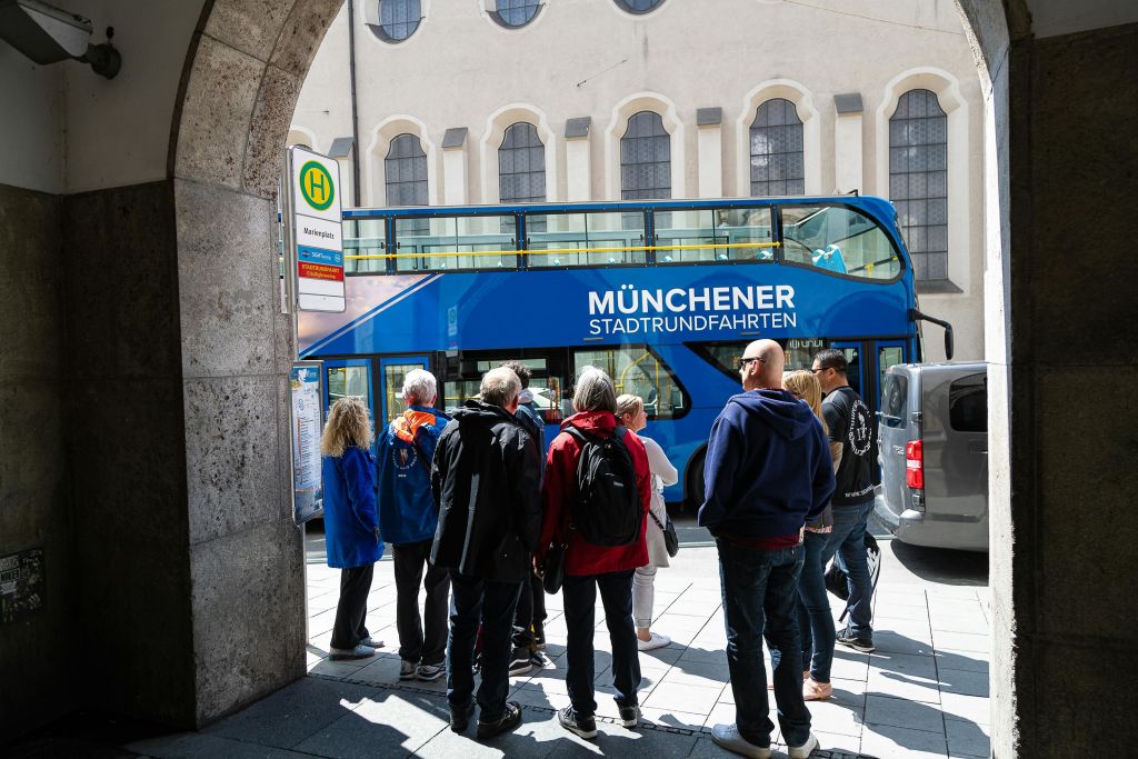 도심의 '뮌헨 슈타트룬트파르텐 (Münchener Stadtrundfahrten)' 이라는 이름의 파란색 2층 투어 버스 근처에 서 있는 한 무리의 사람들.